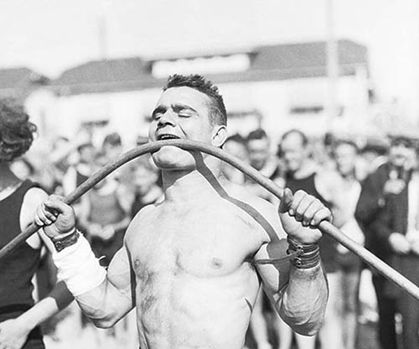 Image of strength feats athlete bending a metal bar held in his mouth